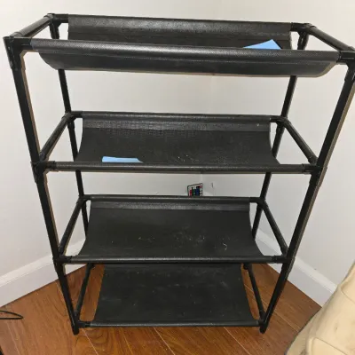 Sale Photo Thumbnail #200: Black lightweight 4-tier shoe or storage rack featuring tubular metal or plastic frame construction with fabric shelf slings. Shows minor wear and dust from use.