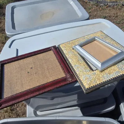 Sale Photo Thumbnail #109: Collection of various decorative wall artwork and photo frames including gilt-finished framed prints, ornate silver-tone and bronze-style tabletop frames, woven rattan/wicker style sunburst accent mirrors, and mosaic/textured picture frames. Plastic stora
