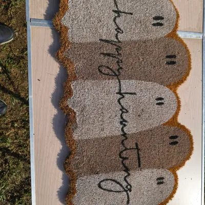 Sale Photo Thumbnail #180: A ghost-shaped Halloween coir doormat featuring script lettering that reads "happy haunting" across multiple neutral-toned ghost figures. Designed for entryway or porch use. Appears to be in unused or lightly used condition.