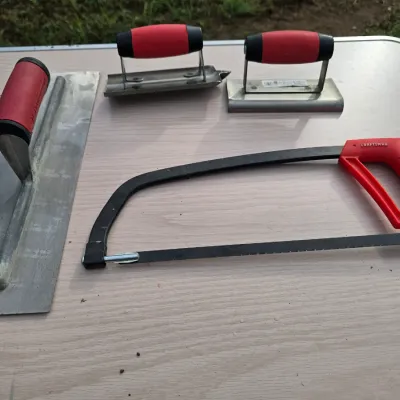 Sale Photo Thumbnail #162: A 4-piece lot of hand tools including a Craftsman hacksaw with a red handle and blade attached, a long metal finishing trowel (Marshalltown/QLT), a concrete groover tool, and an edger. All feature comfortable red and black or red handles. Condition is goo
