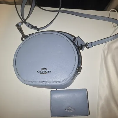 Sale Photo Thumbnail #89: light blue leather set including a round canteen-style crossbody handbag and a matching bi-fold cardholder wallet. The bag features dark silver-tone/gunmetal hardware, an adjustable leather strap, top zip closure, and the iconic metallic Coach logo on the