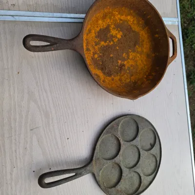 Sale Photo Thumbnail #168: Lot of two cast iron cookware items. Includes a Lodge cast iron skillet with dual pour spouts and a helper handle, alongside a 7-socket silver dollar pancake / Swedish plett pan featuring a flower design on the underside. Both pieces show surface rust and