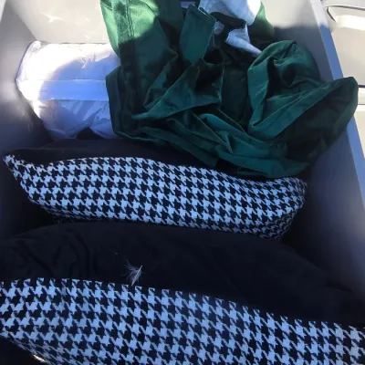 Sale Photo Thumbnail #115: Storage tub containing a collection of household linens and decor items, including two black and white houndstooth pattern throw pillows with solid black backs, a green velvet-like drape or fabric, and packaged white sheers. Plastic storage container with