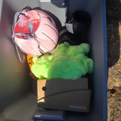 Sale Photo Thumbnail #117: A plastic tote filled with modern Halloween decorations and accessories. Includes a pink jack-o'-lantern bucket with tulle and bow accents, mini cauldrons, decorative fabric ghost favors or ornaments, green faux cobweb fluff, and additional small items. P