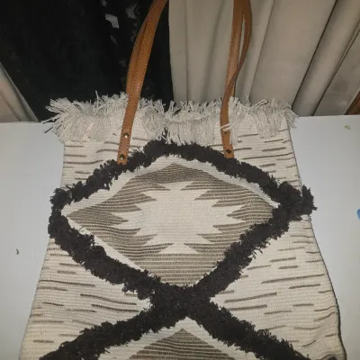 Sale Photo Thumbnail #97: Boho style woven cloth tote bag featuring a geometric Southwestern diamond pattern, dark brown fringed accent trim, fringe along the top edge, and faux leather shoulder straps with metal studs. Gently used condition with no visible tears or major stains.