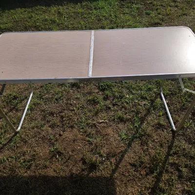 Sale Photo Thumbnail #181: A lightweight folding camp or utility table featuring a faux woodgrain laminate top and a durable tubular aluminum frame and legs. Folds in half for easy transport and storage. Appears to be in good, functional condition with light surface wear from use.