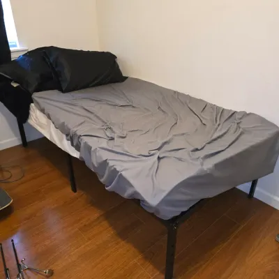 Sale Photo Thumbnail #194: Metal platform bed frame with mattress, grey fitted sheet, and black pillows. Appears to be in functional, used condition. Modern design with open space underneath for storage.