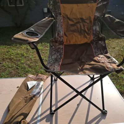 Sale Photo Thumbnail #179: Folding portable outdoor camp chair featuring a camouflage and tan fabric design with orange trim. Includes armrests with a integrated mesh cup holder and a matching carrying bag with shoulder strap. Shows light wear from outdoor use; overall in good usab