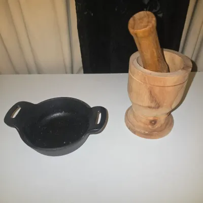 Sale Photo Thumbnail #85: A two-piece kitchen prep lot including a carved wooden mortar and pestle set (Pilón) along with a small double-handled black cast iron serving dish/skillet. Both items are in good pre-owned condition with light signs of use.