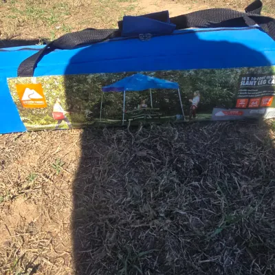 Sale Photo Thumbnail #136: An Ozark Trail 10 ft x 10 ft instant slant leg canopy housed inside a blue zippered carrying storage bag with handles. Features 64 sq. ft. of shade coverage, UPF 50+ protection, and a durable steel frame. Pre-owned condition, stored in original travel bag