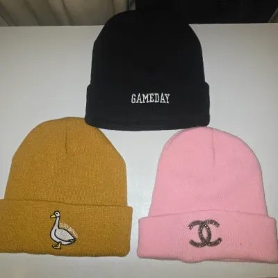 Sale Photo Thumbnail #106: A group of three cuffed knit beanie hats in various colors and embroidered designs: one black beanie embroidered with 'GAMEDAY', one mustard yellow beanie with a 'silly goose' graphic patch, and one pink beanie with an interlocking CC emblem. All appear t