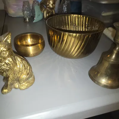 Sale Photo Thumbnail #78: A collection of brass decorative items including a detailed sitting cat figurine, a small polished brass bowl, a large ribbed/swirled brass bowl, and a brass candlestick holder. Items show light surface wear and minor tarnishing consistent with age and us