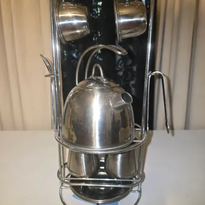 Sale Photo Thumbnail #69: A polished stainless steel mini tea or coffee set featuring a teapot with lid, cups, saucers, and utensils, all organized on a multi-tier chrome-finished metal wire display stand. Light surface wear and fingerprints visible on the polished finish, overall