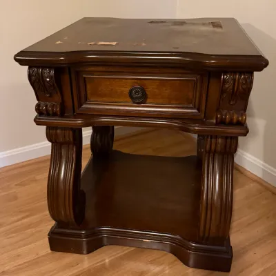Sale Photo Thumbnail #12: Traditional dark wood end table featuring a single drawer with round brass hardware, carved acanthus leaf details, and a lower shelf. The top surface shows noticeable finish wear and peeling veneer.