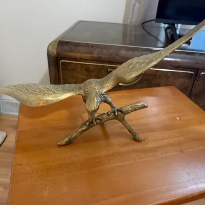 Sale Photo Thumbnail #366: Vintage solid brass eagle figurine depicted in flight, perching on a wooden-style branch, displayed on a wooden table.