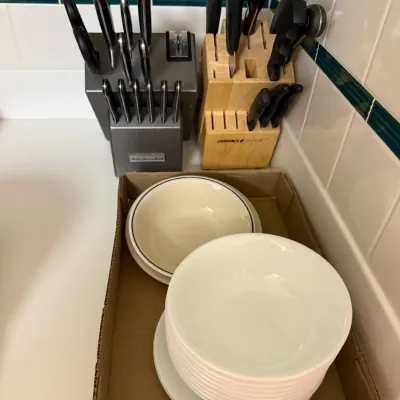 Sale Photo Thumbnail #294: Two wooden and metal knife blocks filled with cutlery, positioned behind stacked ceramic bowls in a cardboard tray.