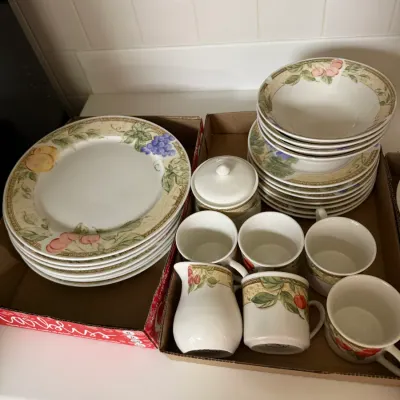 Sale Photo Thumbnail #292: A multi-piece ceramic dinnerware set featuring a fruit motif border, including plates, bowls, mugs, a cream pitcher, and a covered sugar bowl.