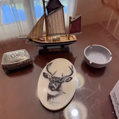 Sale Photo Thumbnail #256: Collection of tabletop decor including a miniature model sailboat, an oval scrimshaw-style plaque depicting a buck, an embossed metal trinket box, and a ceramic ashtray.