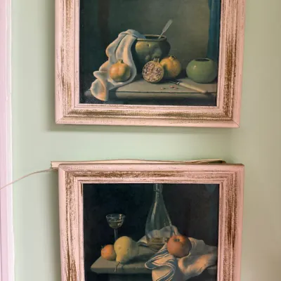 Sale Photo Thumbnail #296: Pair of framed still life art prints depicting fruit, glassware, and tableware, housed in matching distressed whitewashed wooden frames.