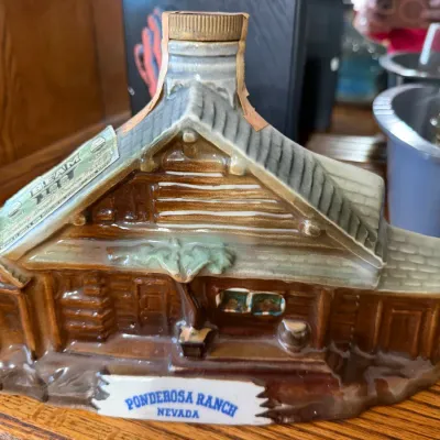 Sale Photo Thumbnail #329: Vintage Jim Beam ceramic decanter shaped like the Ponderosa Ranch log cabin, featuring original tax stamp and labels.