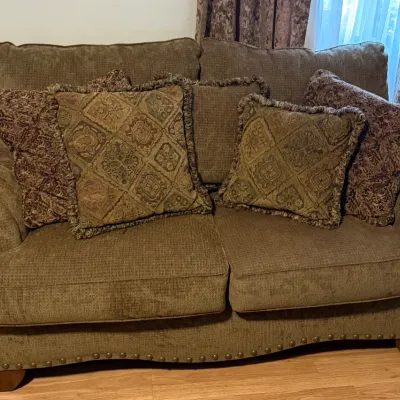 Sale Photo Thumbnail #79: Plush, olive green upholstered loveseat featuring rolled arms, brass nailhead trim along the base, and an assortment of patterned accent pillows.