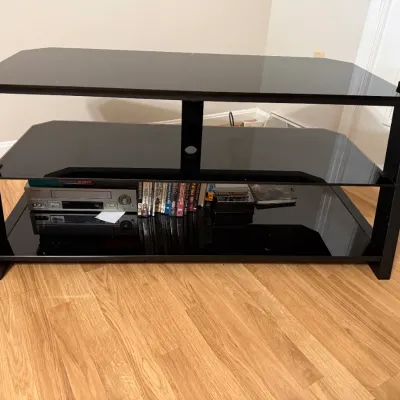 Sale Photo Thumbnail #32: Modern black metal and tempered glass three-tier media console, holding a silver VCR and media tapes on the lower shelf.
