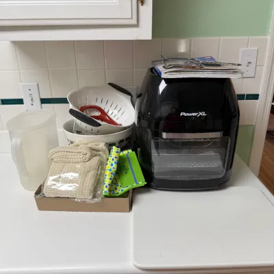 Sale Photo Thumbnail #150: Black PowerXL air fryer sitting on a kitchen counter next to colanders, a plastic pitcher, folded towels, and a cutting board.