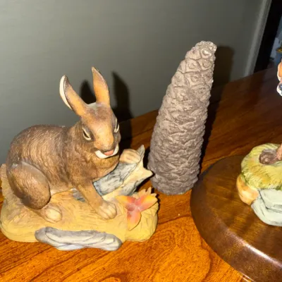 Sale Photo Thumbnail #277: Hand-painted porcelain rabbit figurine depicted on a log, displayed alongside a decorative pinecone sculpture on a wooden surface.