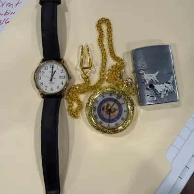 Sale Photo Thumbnail #418: Collection of men's accessories including a black leather strap Timex wrist watch, a ornate gold-tone pocket watch with chain featuring the Great Seal of the United States, and a vintage Zippo-style lighter decorated with hunting dogs.