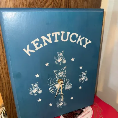 Sale Photo Thumbnail #270: Vintage blue wooden Kentucky Wildcats wall clock featuring wildcat mascot graphics and brass-tone clock hands.