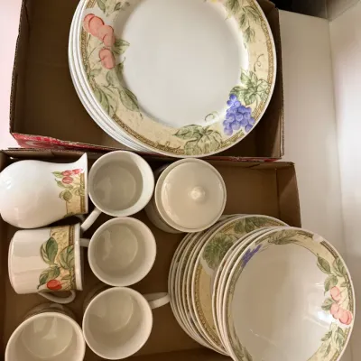 Sale Photo Thumbnail #293: Set of ceramic dinnerware featuring a fruit motif border, including dinner plates, bowls, teacups, a pitcher, and a sugar bowl stored in cardboard boxes.