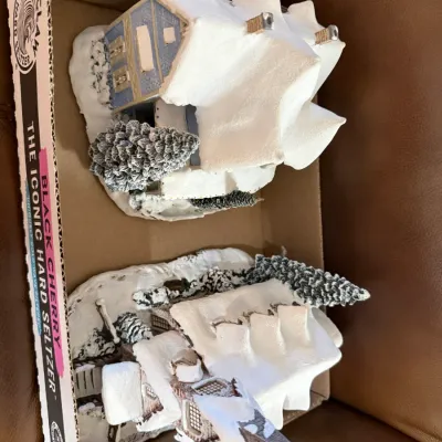 Sale Photo Thumbnail #348: Pair of snow-covered miniature Christmas village buildings, including a house and a church with a steeple, stored in a cardboard box.