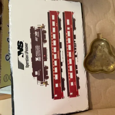 Sale Photo Thumbnail #264: Norfolk Southern Operation Lifesaver printed slate plaque depicting a train, shown alongside a small, pear-shaped brass ash tray in a cardboard box.