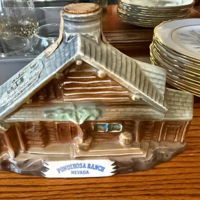 Sale Photo Thumbnail #325: Vintage ceramic decanter shaped like a log cabin, featuring 'Ponderosa Ranch Nevada' lettering on the base and an original paper whiskey label on the roof.