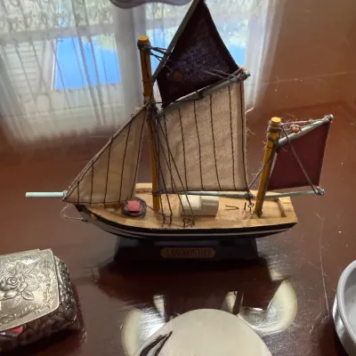Sale Photo Thumbnail #258: Handcrafted miniature wooden model ship titled 'Langoustier', featuring fabric sails, detailed rigging, and a black and white hull resting on a wooden display base.
