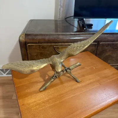Sale Photo Thumbnail #365: Vintage brass eagle sculpture with outstretched wings perched on a branch, displayed on a wooden folding table.