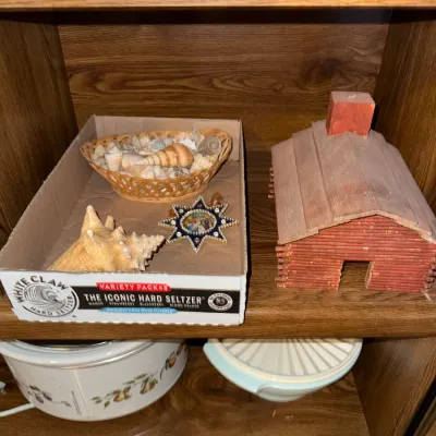 Sale Photo Thumbnail #284: Collection of decorative items including a miniature wooden log cabin house, a basket of seashells, an ornate star-shaped ornament, and kitchenware displayed on wooden shelving.