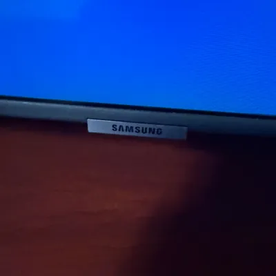 Sale Photo Thumbnail #126: Close-up of a Samsung television bezel showing the brand logo badge on a lower dark bezel, powered on with a blue screen display.