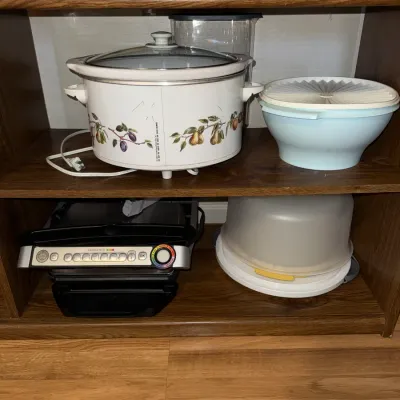 Sale Photo Thumbnail #287: Shelved kitchen appliances including a Rival Crock-Pot with fruit motif, a T-fal OptiGrill, a vintage light blue Tupperware bowl, and a plastic cake carrier.