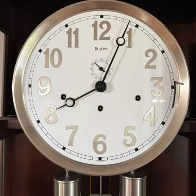 Sale Photo Thumbnail #2: Bulova grandfather clock dial featuring a silver-tone bezel, raised numerals, second sub-dial, and visible chimes and weights.