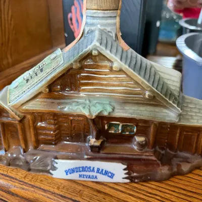 Sale Photo Thumbnail #328: Vintage Jim Beam ceramic decanter shaped like the Ponderosa Ranch log cabin, featuring original tax stamp and Nevada souvenir banner.