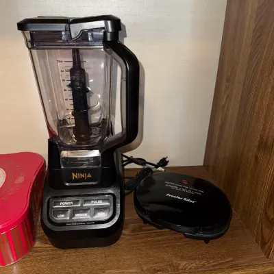 Sale Photo Thumbnail #269: Black Ninja Professional 1000W countertop blender with plastic pitcher alongside a black Proctor Silex electric appliance on a wooden shelf.