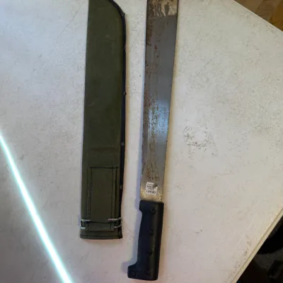 Sale Photo Thumbnail #184: Military-style machete with a black composite handle, displaying surface rust, accompanied by an olive green canvas sheath.