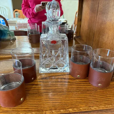 Sale Photo Thumbnail #333: Vintage cut glass decanter with faceted stopper and hanging silver-tone label, accompanied by four rocks glasses featuring snap-on leather wraps.