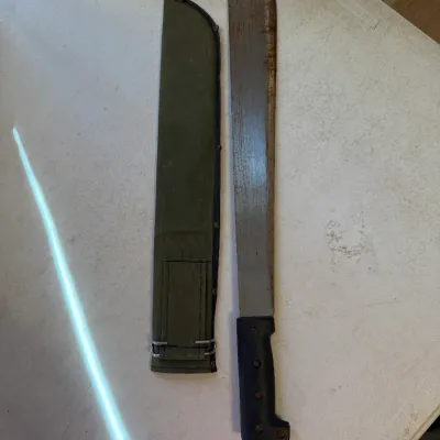 Sale Photo Thumbnail #183: Vintage military-style machete with a black textured handle and a long steel blade, accompanied by an olive green canvas sheath.
