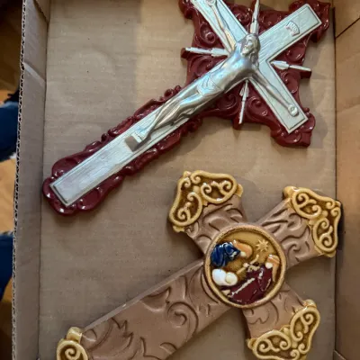 Sale Photo Thumbnail #224: Two decorative religious wall crosses resting inside a cardboard box, including a silver-tone crucifix on a red base and a ceramic cross with a central nativity scene detail.