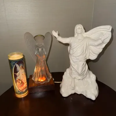 Sale Photo Thumbnail #230: Collection of religious decor featuring a white ceramic Jesus figurine, an illuminated glass angel lamp on a wooden base, and a tall prayer candle.