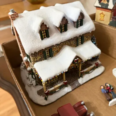 Sale Photo Thumbnail #356: Miniature Christmas village stone house collectible with snow-covered roof, dormant windows, and accompanying red vintage toy car and figurines.