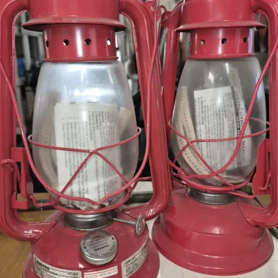 Pair of modern red painted kerosene hurricane lanterns with glass globes, wicks, and instruction manuals stored inside.
