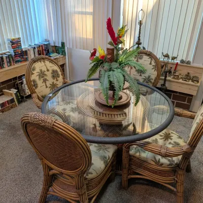 Tropical-style rattan dining set with a round glass-top table and four upholstered armchairs featuring palm tree print fabric.